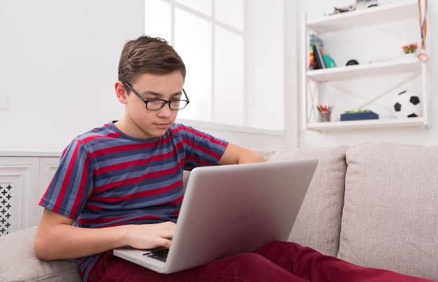 Serious boy with laptop computer on sofa at home. Child surfing the web, typing on keyboard