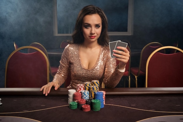 반짝이는 금색 드레스를 입은 진지한 브루네트 여성이 연기 속에서 카지노 테이블에 앉아 포커를 하고 있습니다. 그녀는 큰 승리를 기다리고 카메라를보고 내기를하고 있습니다. 돈을 위한 도박. 게임