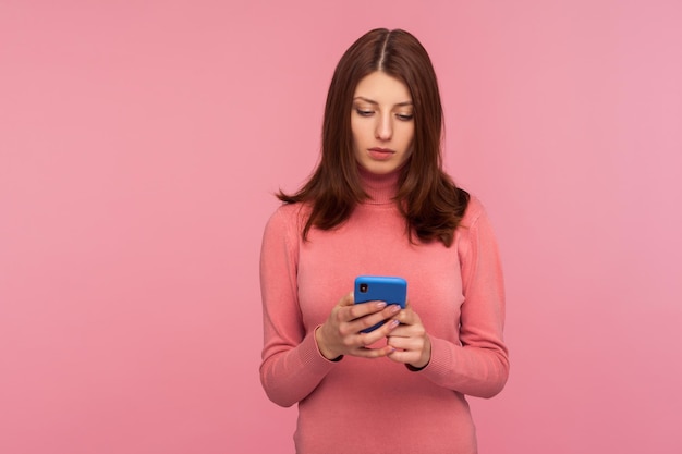 Serious brunette woman holding smartphone in hands chatting with friends or making food order online services Indoor studio shot isolated on pink background