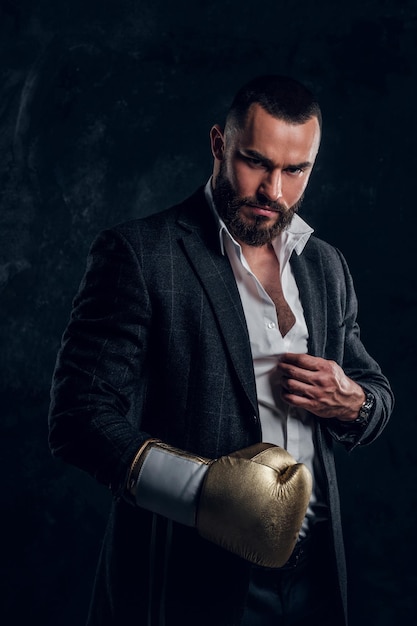 Serious brutal man in suit and golden boxing glove is posing for photographer at dark photo studio.