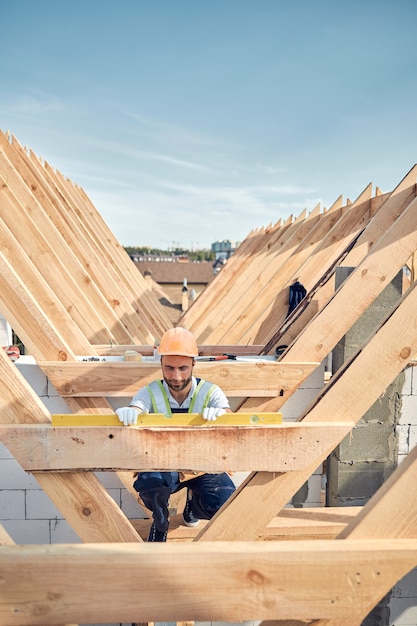 Photo serious builder in a hardhat kneeling near a wooden bar and using a level instrument