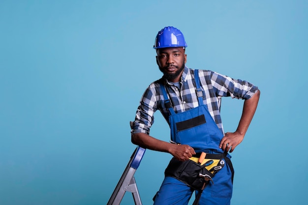 Серьезный строитель, опирающийся на лестницу в комбинезоне и защитном шлеме, смотрит в камеру на голубом фоне. Студия снята с концепцией строительной отрасли.