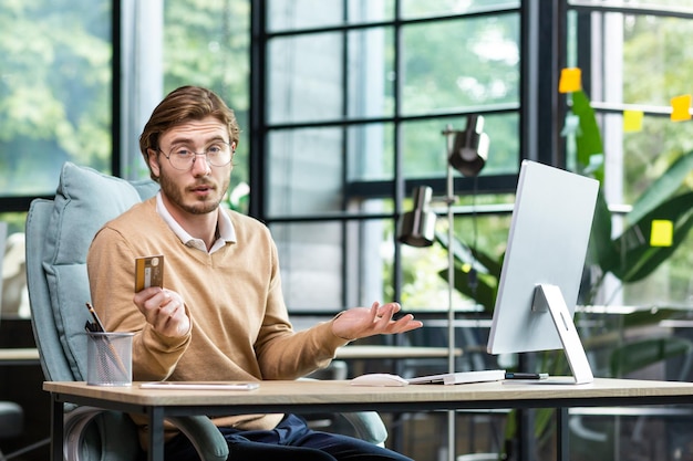 Serious business man sitting at desk in home office holding credit card worried about debt and