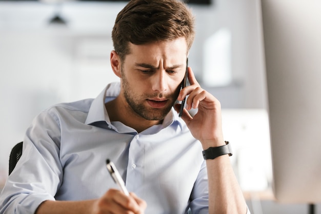 Serious business man talking by smartphone and writing something while sitting by the table in office