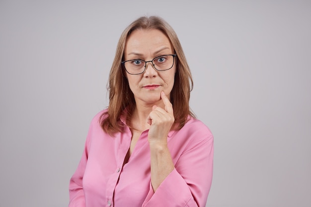 Serious business woman in glasses posing, looking at the camera, hand at the chin. Photo on gray background with copy space.