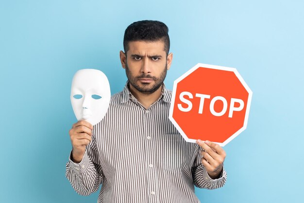 Serious businessman holding white mask with unknown face and red traffic sign looking at camera