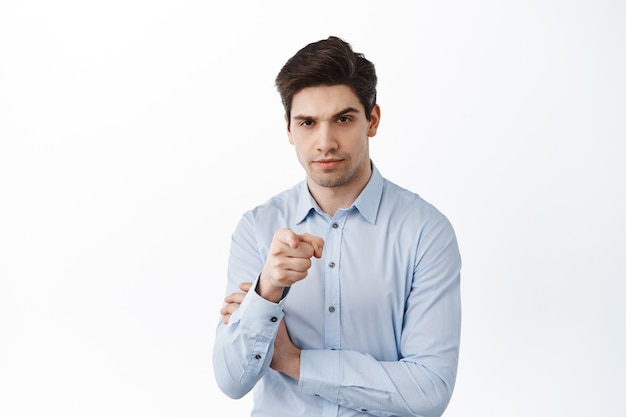 Serious businessman pointing at front, talking to you, choosing employee, inviting to work in his company, standing over white wall