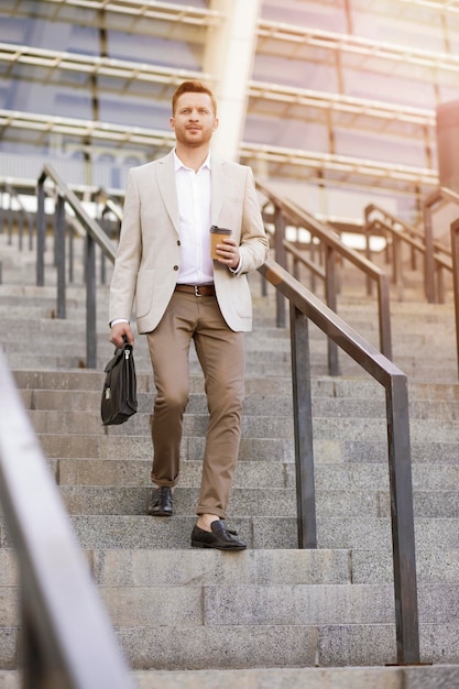Серьезный бизнесмен быстро спускается по лестнице с чемоданом и чашкой кофе в руках