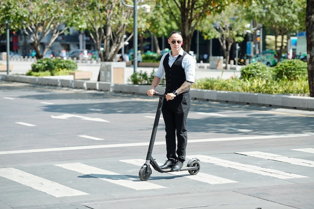 Serious businessman in sunglasses riding electric scooter on crossroads