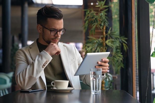 serious businessman wearing suit and eyewear using digital tablet sitting at table in a cafe communicating online checking emails distantly working