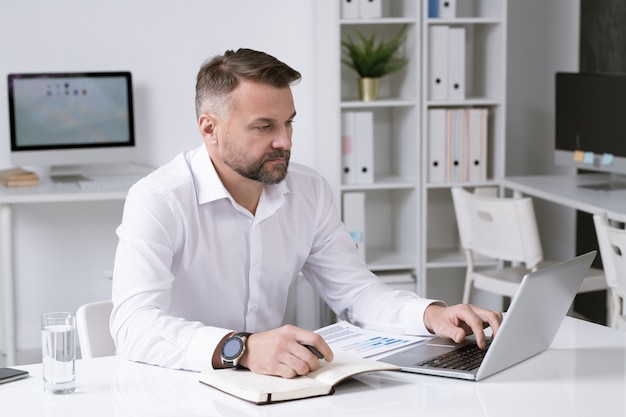 Серьезный бизнесмен в белой рубашке просматривает данные на дисплее ноутбука, сидя за столом в офисе