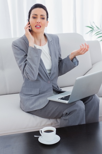 Serious businesswoman calling with her mobile phone and using laptop sitting on sofa 