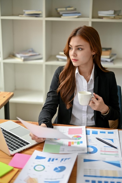 A serious businesswoman sits at her desk with financial reports looking out the window