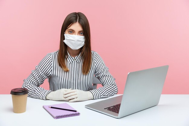 Serious businesswoman sitting safe healthy with hygienic face mask and protective gloves during quarantine, looking at camera. Working at home office in self-isolation, coronavirus outbreak. indoor