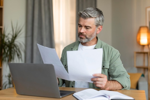 Serious busy old caucasian man manager works with documents sits at table with computer in living room