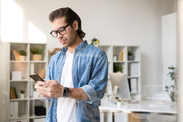 Serious busy young brunette man in glasses checking phone message in own office