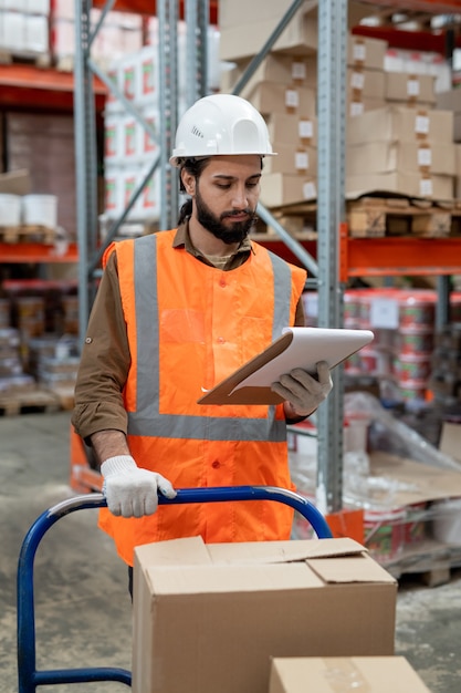 Serious busy young mixed race warehouse worker in hardhat reading consignment note while examining item placement