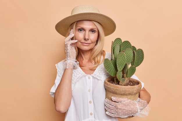 Serious calm blonde European woman looks directly at front wears fashionable outfit holds potted cactus takes care of houseplant isolated over brown wall