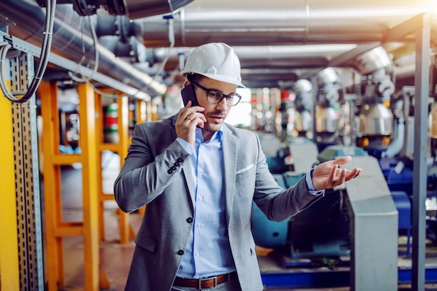 Uomo d'affari caucasico serio dentro con il casco sulla testa che ha telefonata mentre stando nella centrale elettrica.