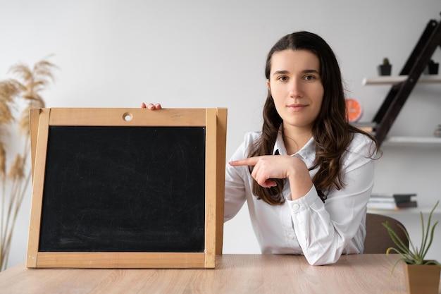 Serious caucasian teacher sitting in the classroom points his\
finger at the chalk board with space for text back to school\
concept caucasian millennial girl in the office holds a blackboard\
student