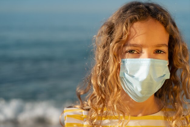 Serious child wearing medical mask outdoor against blue sky wall.