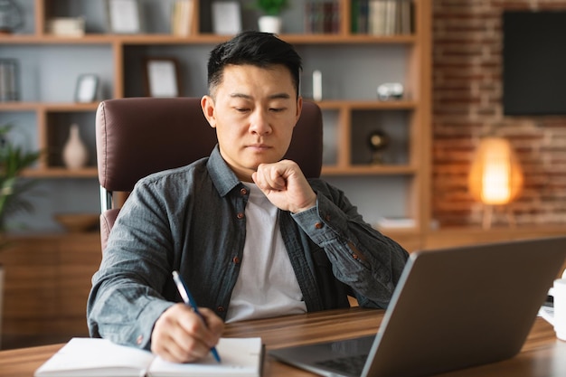 Serious concentrated adult asian man makes notes analyzes data for investment at workplace with laptop