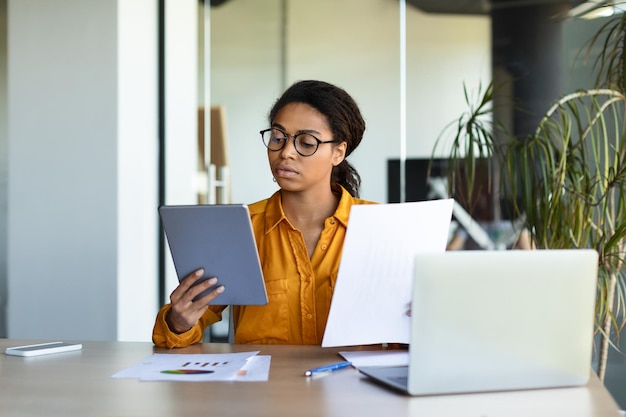 Serious concentrated black businesswoman analyzing data in documents and tablet sitting at workplace in office interior