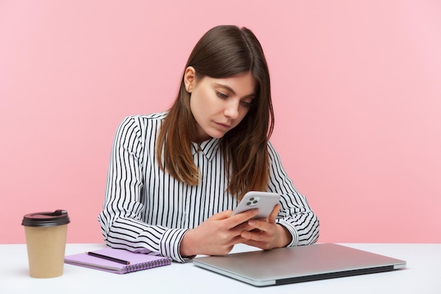 Серьезная сосредоточенная деловая женщина в полосатой рубашке ведет деловую переписку на своем смартфоне, сидя на рабочем месте и ведя блог. Внутренний студийный снимок изолирован на розовом фоне