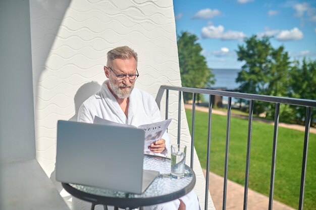 Serious concentrated male in eyeglasses and the white bathrobe sitting on the balcony with a newspaper in the hands