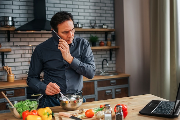 Serious concentrated man stand at kitchen table and look at laptop screen. He talk on phone and blend eggs in bowl. Colorful vegetables lying on desk.