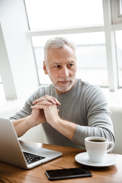 Un uomo d'affari senior maturo concentrato serio si siede in caffè facendo uso del computer portatile.