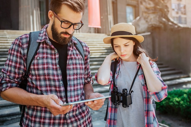 Serious and concentrated tourists stand at steps. He holds tablet in hands. They both look at it. Young woman hold lace from binoculars. Travellsers are very thoughtful.