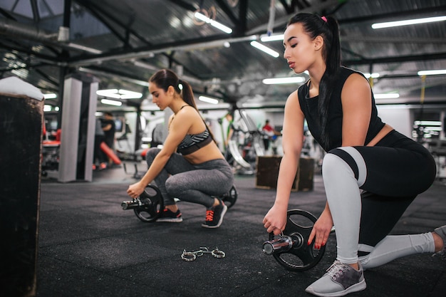 Serious an concentrated young asian woman stand on one knee and look down. She put some weight to barbell. Her european friend repeat actions after her. They are in gym.