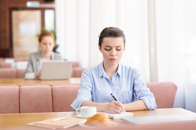 Serious concentrated young businesswoman in shirt sitting at table in modern cafe and making notes on business plan