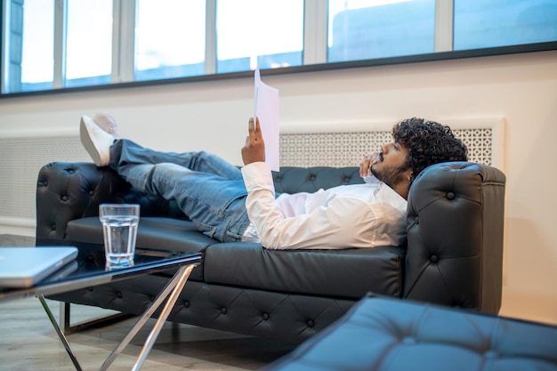 Serious concentrated young Indian male entrepreneur lying on the leather sofa and perusing business papers