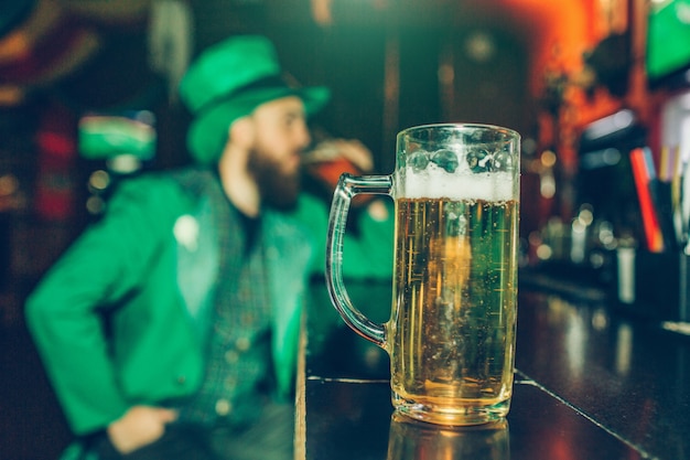 Photo serious concentrated young man in st. patrick's suit sit at bar counter in pub alone. mug of beer stand in front of him closer to camera