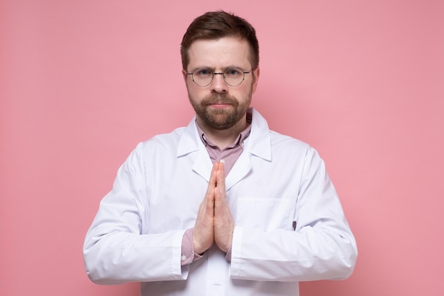 Serious confident doctor in white coat folded hands in supplication palms together and calmly looks