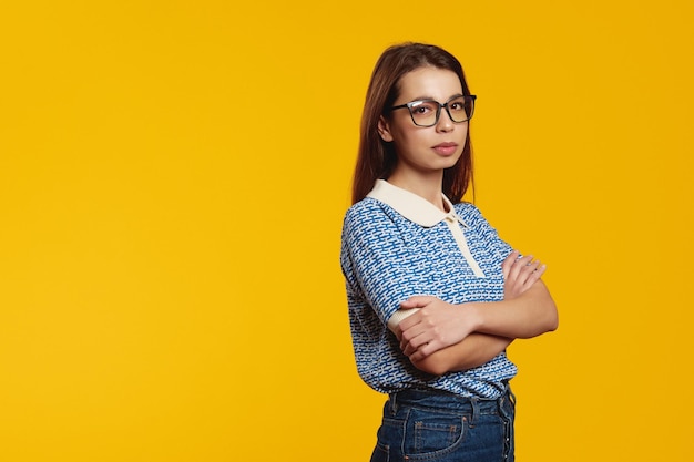 Serious confident girl with eyeglasses looking at camera with crossed arms isolated over yellow
