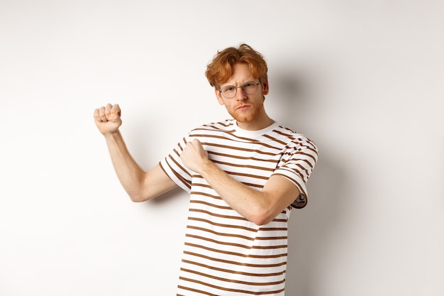 Serious and confident redhead man in glasses raising fists, ready for fight, staring angry at camera while shadow boxing, standing over white background