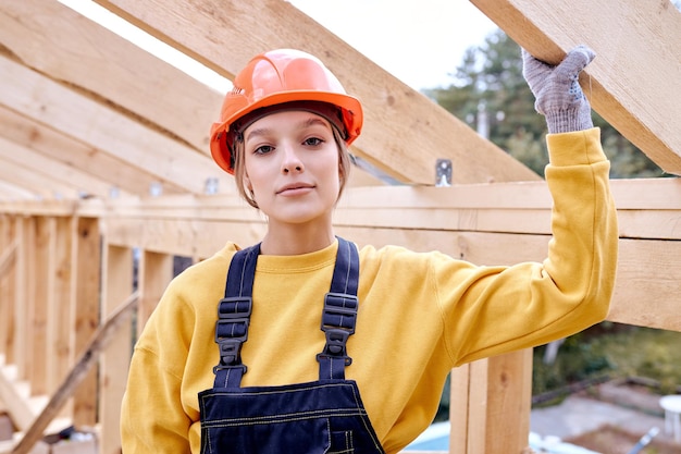 新しい住宅建設の家のフレーミングの深刻なコンストラクターエンジニアの女性。木造建築現場。オレンジ色のヘルメットのヘルメットとカメラでポーズをとって制服を着た魅力的な白人女性