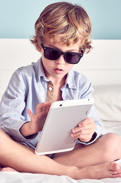 Serious cool boy in stylish sunglasses sitting with crossed legs and surfing tablet on bed in bedroom