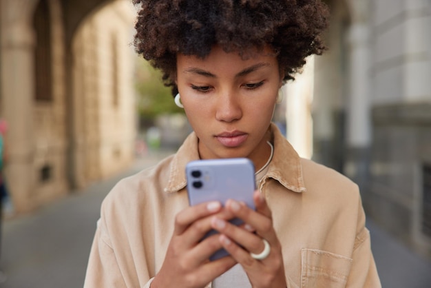 真面目な縮れ毛の女性がスマートフォンでテキストメッセージを送信検索街の通り検索正しい方向使用アプリケーション着用カジュアルジャケット人々現代のテクノロジーとライフスタイルのコンセプト