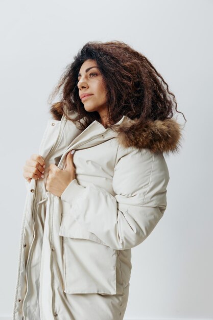 Serious curly pretty latin lady wearing in soft warm beige\
winter coat with fur hood posing isolated looking aside at white\
studio background fashion huge sale new collection concept vertical\
shot