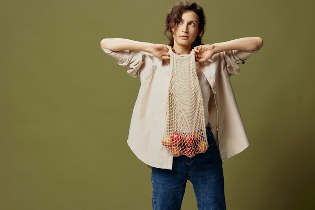 Serious cute curly beautiful lady in linen casual shirt pulls\
string bag with fruit near chest posing isolated on over olive\
green pastel background ecofriendly natural products concept copy\
space