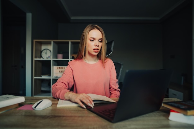 Donna carina seria in maglione rosa che lavora al computer portatile alla scrivania con la faccia seria