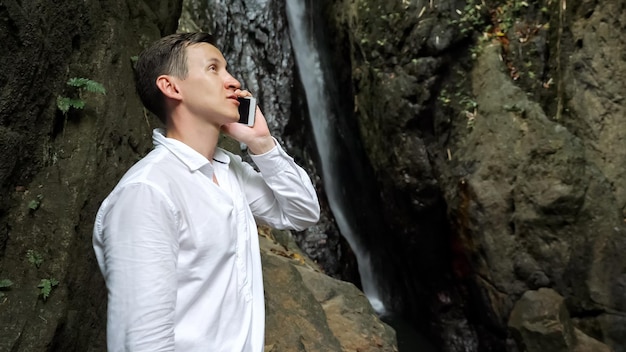 Serious dark haired guy in white shirt holds smartphone in hand and talks on phone standing against tropical waterfall in nature reserve closeup