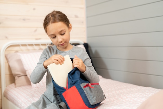 Serious diligent schoolgirl in grey dress putting book into backpack while sitting on bed in bedroom in the morning and going to go to school