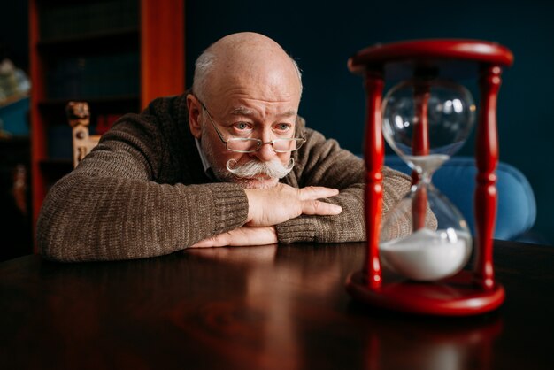 Photo serious elderly man looking on sandglass in home office, time cannot be turned back. mature senior looking on hourglass