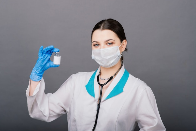 Serious female doctor in a medical mask holding syringe and vaccine isolated on grey