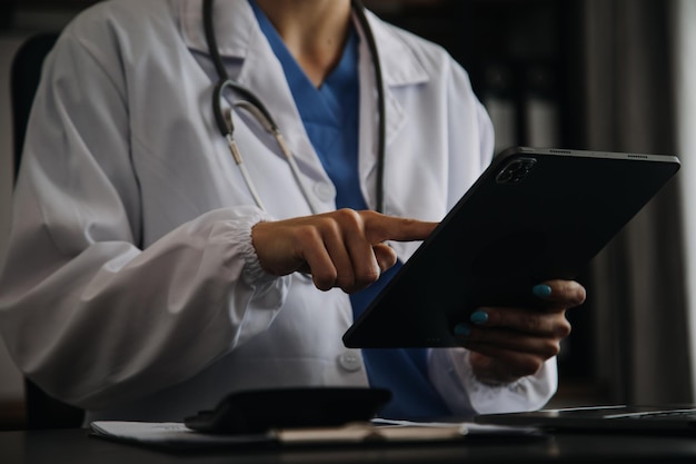 노트북을 사용하는 진지한 여성 의사와 책상에 앉아 있는 의학 저널에 노트를 쓰는 젊은 여성 전문 의료 의사는 흰색 코트를 입고 직장에서 컴퓨터 작업을 하는 청진기를 착용합니다.